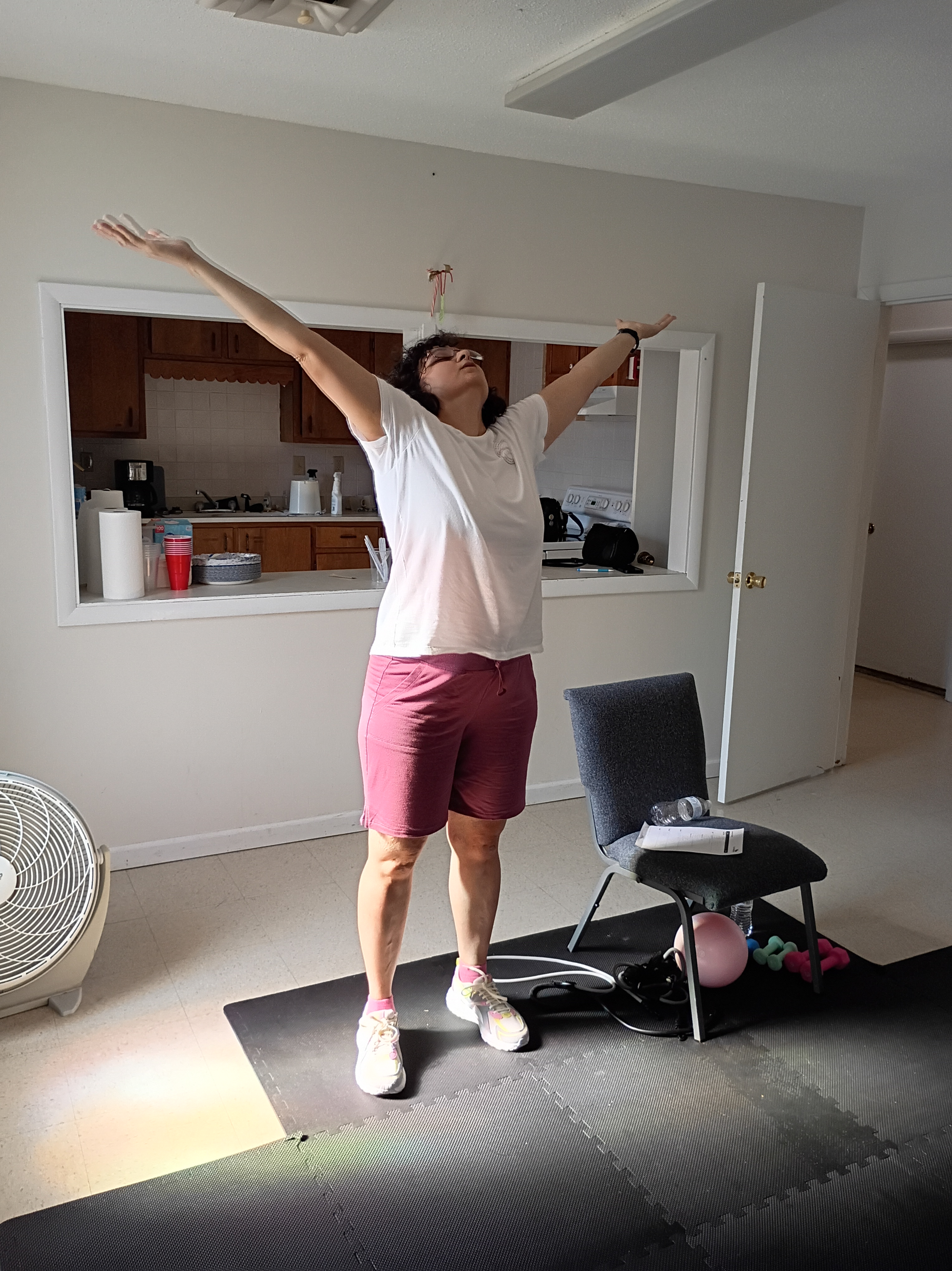 Woman raising her arms in praise. A chair is nearby with fitness equipment on and under it.