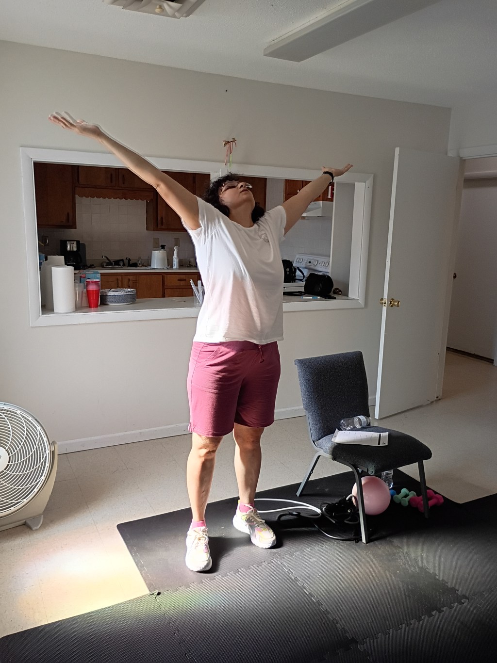 Woman raising her arms in praise. A chair is nearby with fitness equipment on and under it.